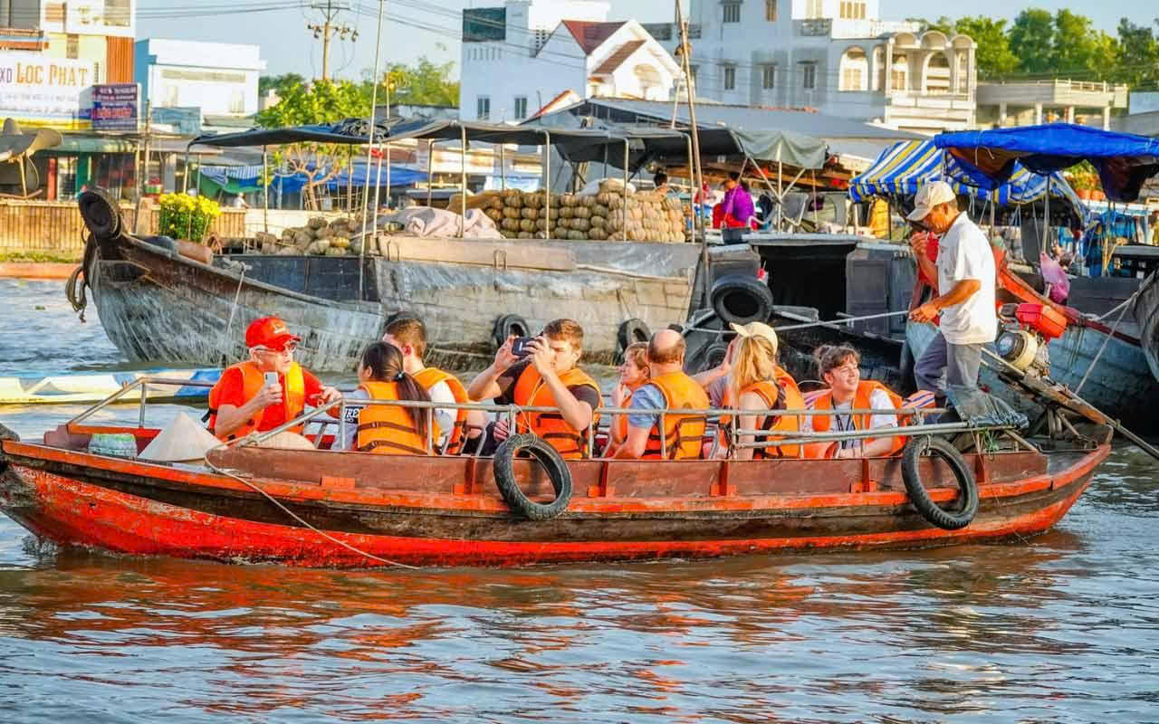 Cette image montre des touristes étrangers portant des gilets de sauvetage lors d'une excursion en barque traditionnelle en bois sur un marché flottant du delta du Mékong, au Vietnam. La barque navigue sur un fleuve animé où des vendeurs locaux proposent fruits et produits agricoles directement depuis leurs embarcations. Ces marchés flottants constituent un élément culturel unique du sud du Vietnam et offrent aux visiteurs un aperçu authentique du mode de vie fluvial et des traditions commerciales des communautés locales.